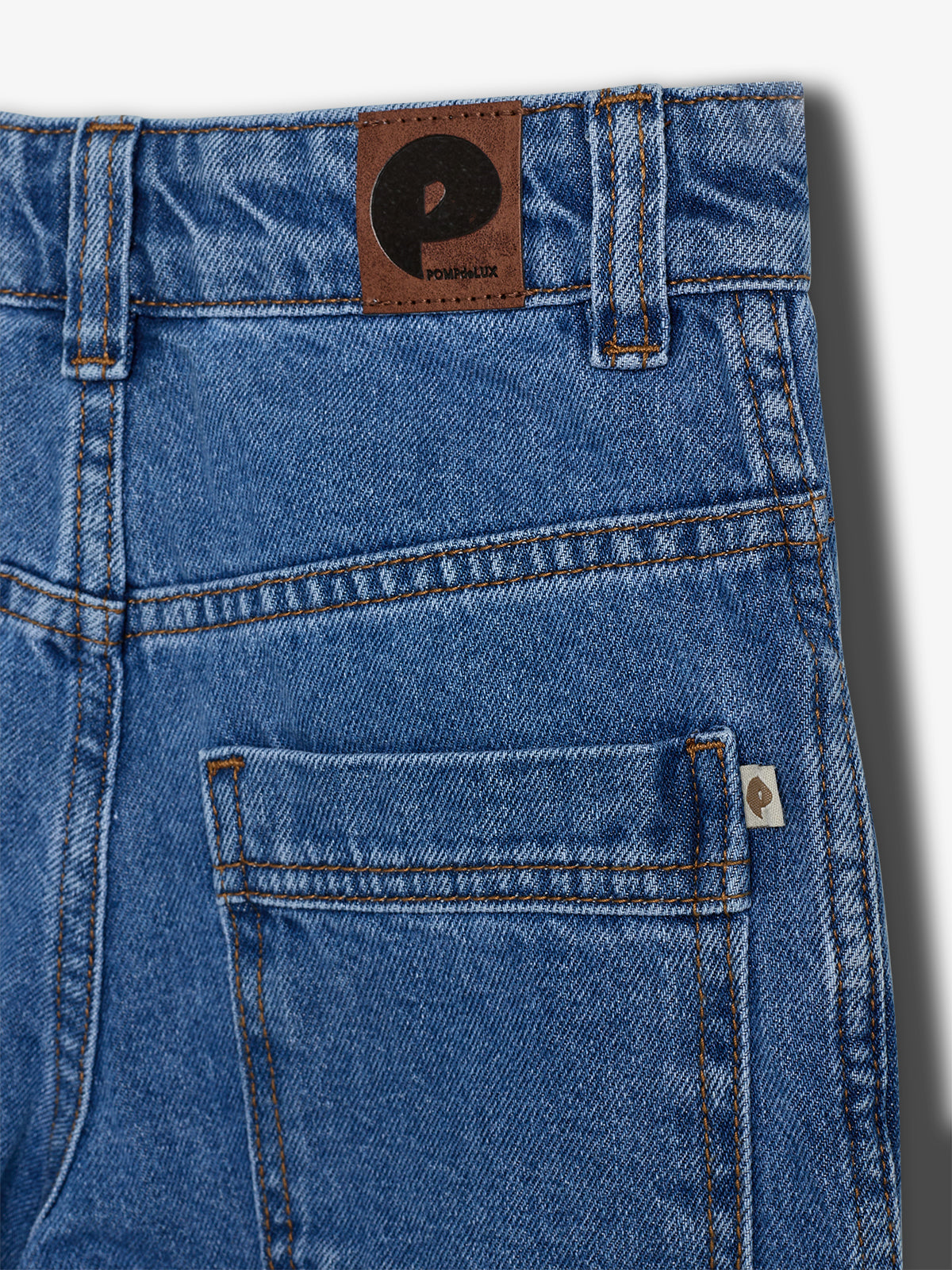 Blue denim jeans with prominent stitching and a brown leather patch featuring a large "P" and "POMPKLUX" text. A small white tag with a "P" is near the pocket.