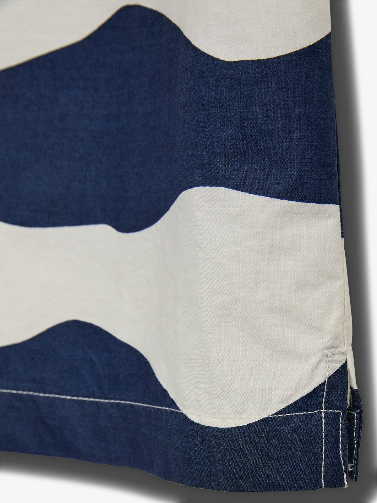 Fabric with wavy blue and white pattern hangs vertically, showcasing detailed stitching near the edge. The background is plain, highlighting the fabric's texture and design.