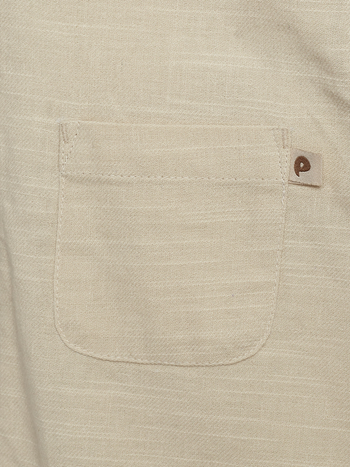 A beige fabric pocket is sewn onto a similar-colored material. A small tag with a brown logo protrudes from the pocket's top right corner.