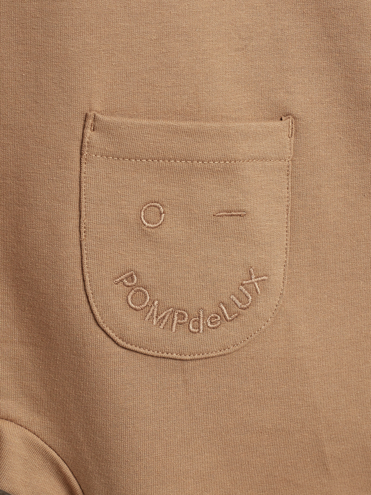 Beige fabric features a pocket with a minimalist face design and the embroidered text "POMPdeLUX." The setting indicates a close-up of clothing material.