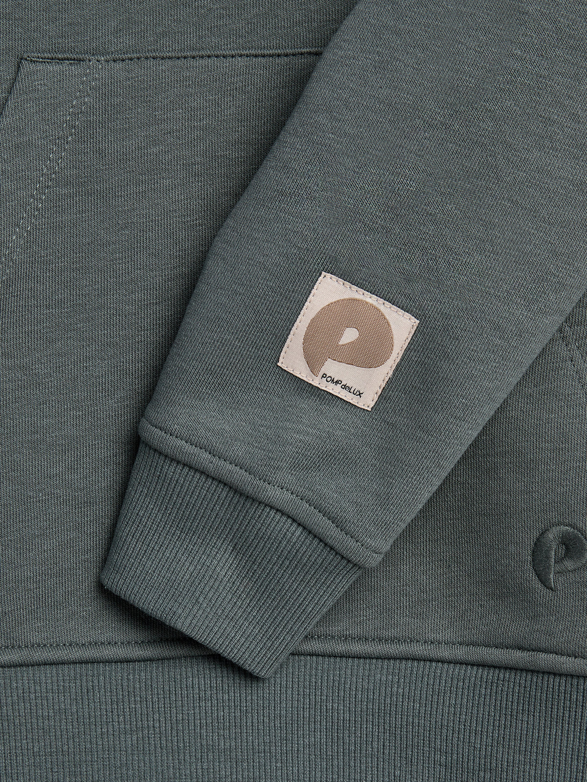 A green sweatshirt sleeve with a patch bearing the letter "P" and text "POP Trading Company," rests over the main body, showing pocket stitching in a neutral setting.