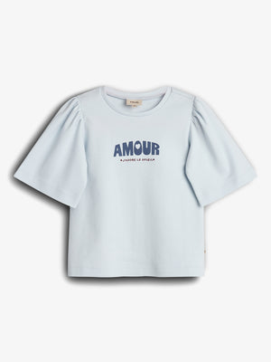A short-sleeved, light blue shirt with puffed sleeves displays the text "AMOUR" in bold, large letters and "J'ADORE LE SOLEIL" beneath. The shirt is on a plain background.
