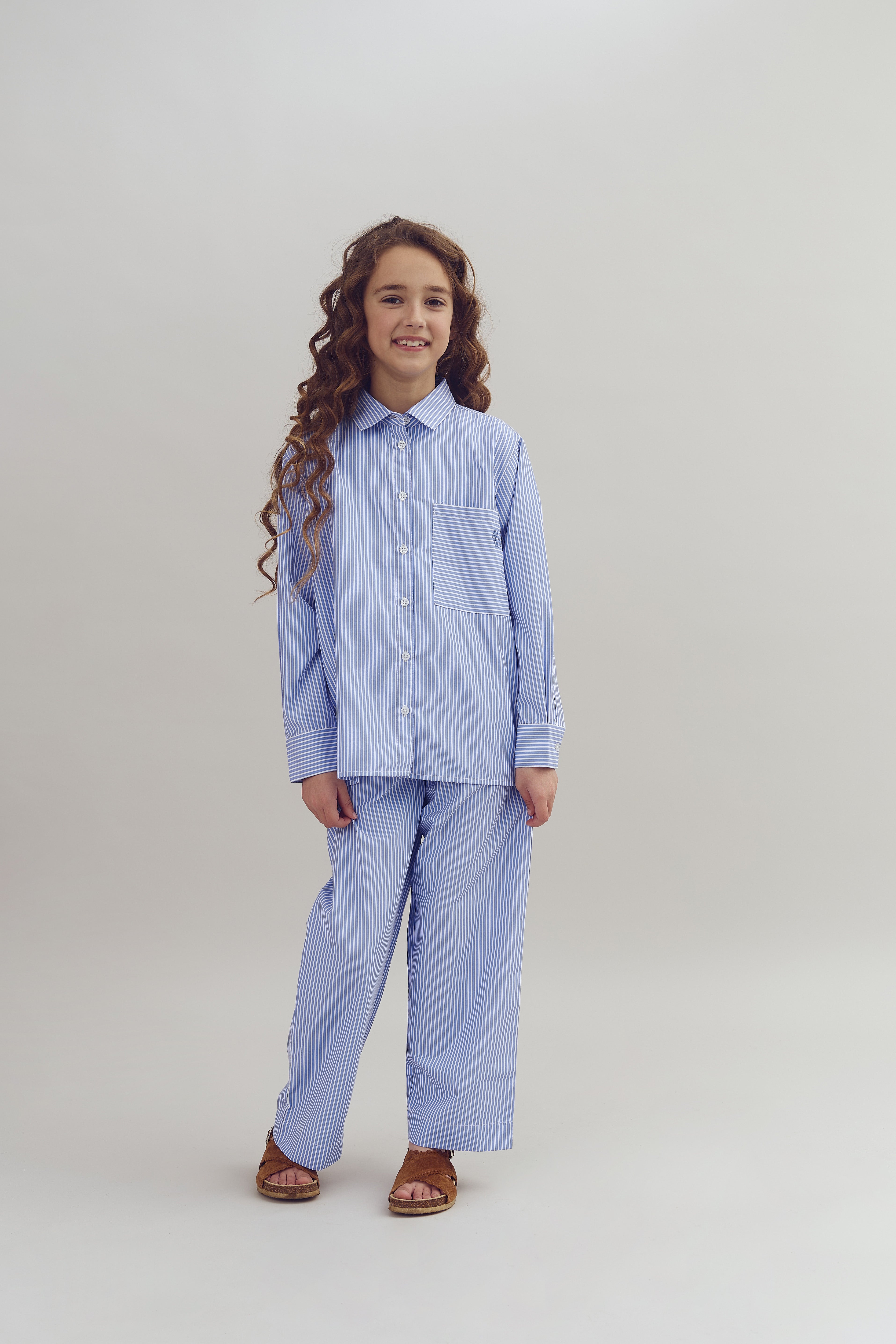 A girl wearing a blue and white striped outfit stands smiling, with loose, curly hair, in a neutral-colored studio setting.