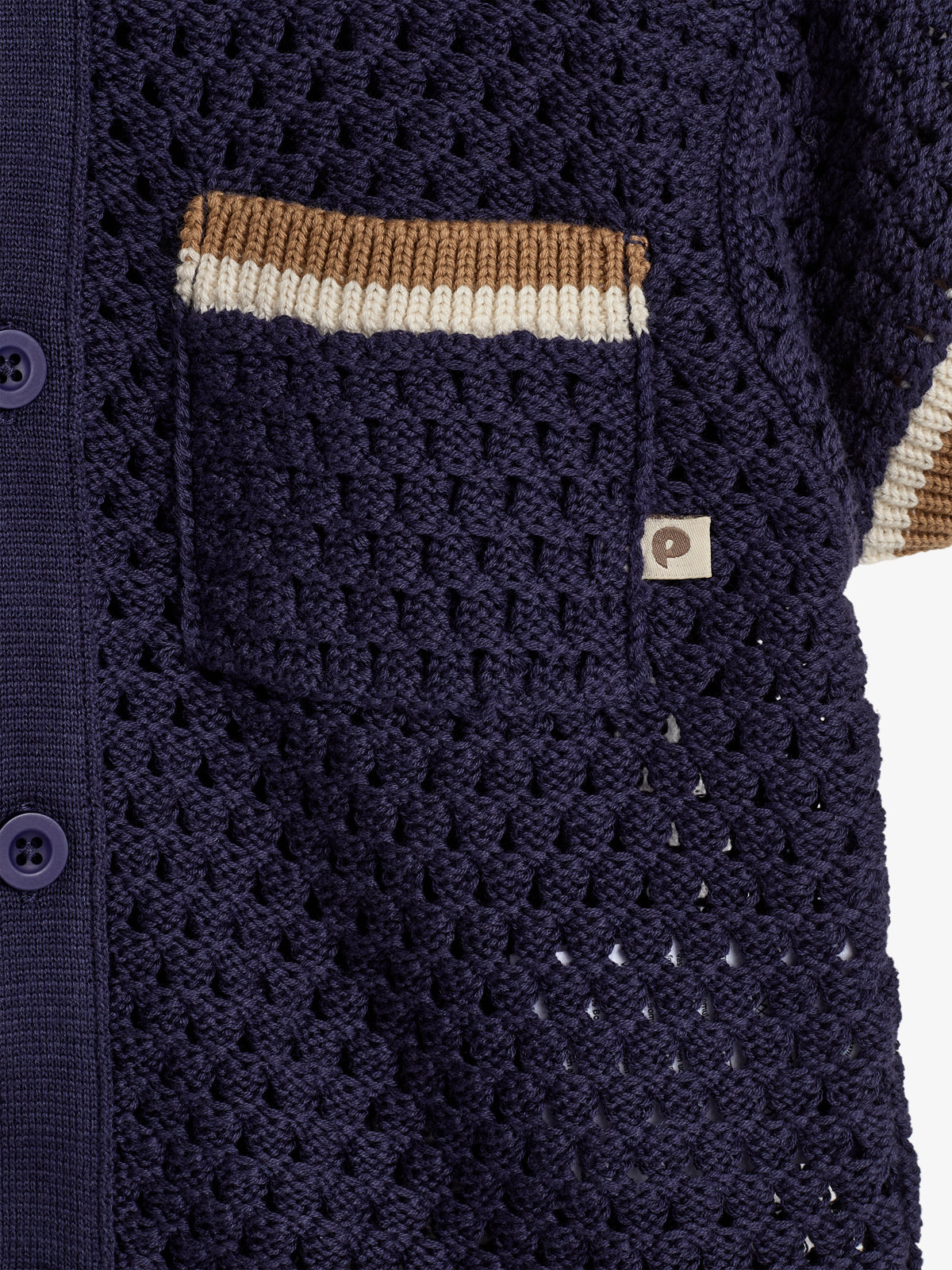A dark blue crocheted fabric features a pocket with brown and white stripes. Four buttons line the edge, and a small tag with a "P" logo is attached.