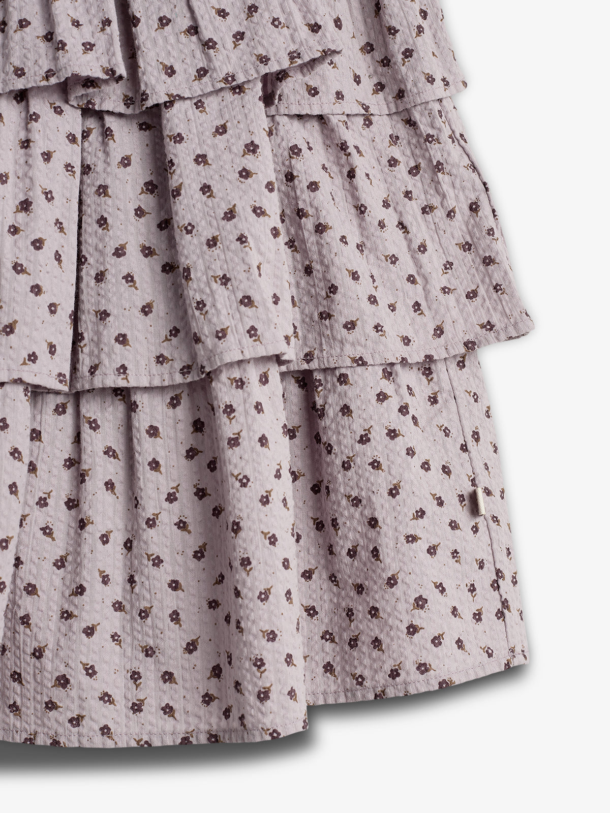 The image shows a light purple, tiered fabric with a small floral pattern. The fabric layers create a textured appearance, suggesting a skirt or dress, set against a plain background.