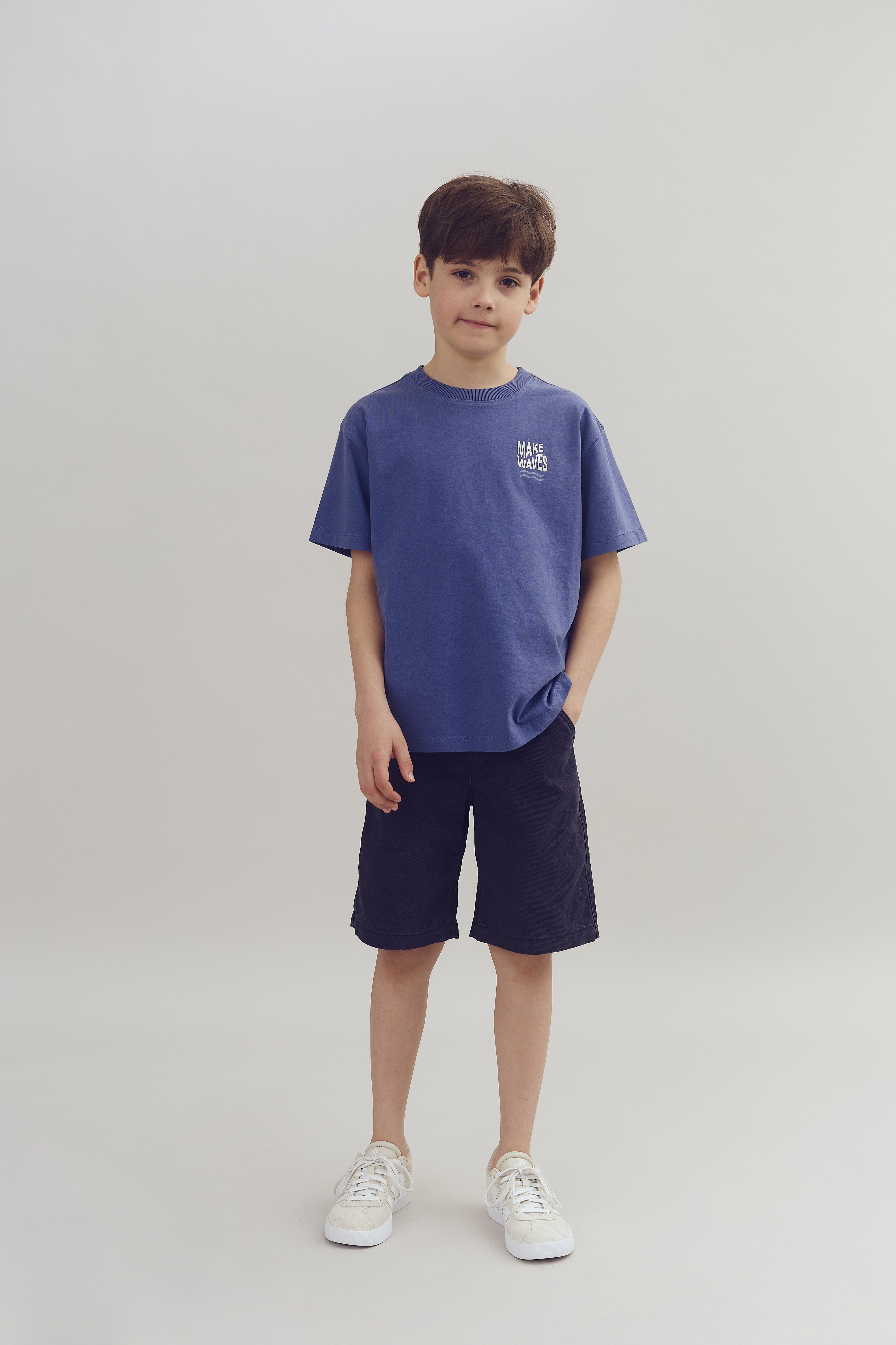 A boy stands casually in a studio, wearing a blue shirt with "MAKE WAVES" on it, dark shorts, and white sneakers. The background is plain and light-colored.