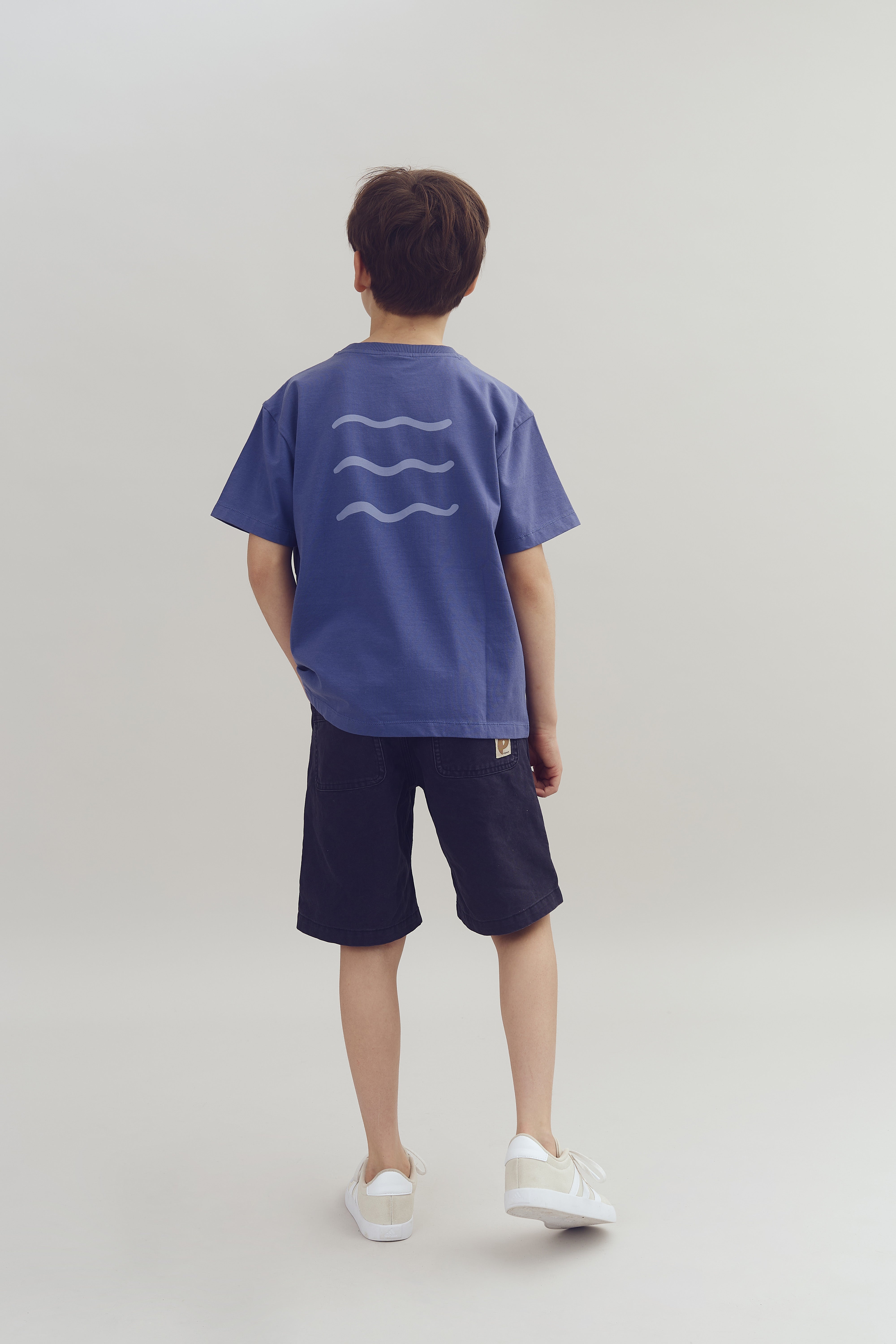 A child wearing a blue t-shirt with wavy line designs stands facing away, dressed in dark shorts and white sneakers, set against a plain, light-colored background.