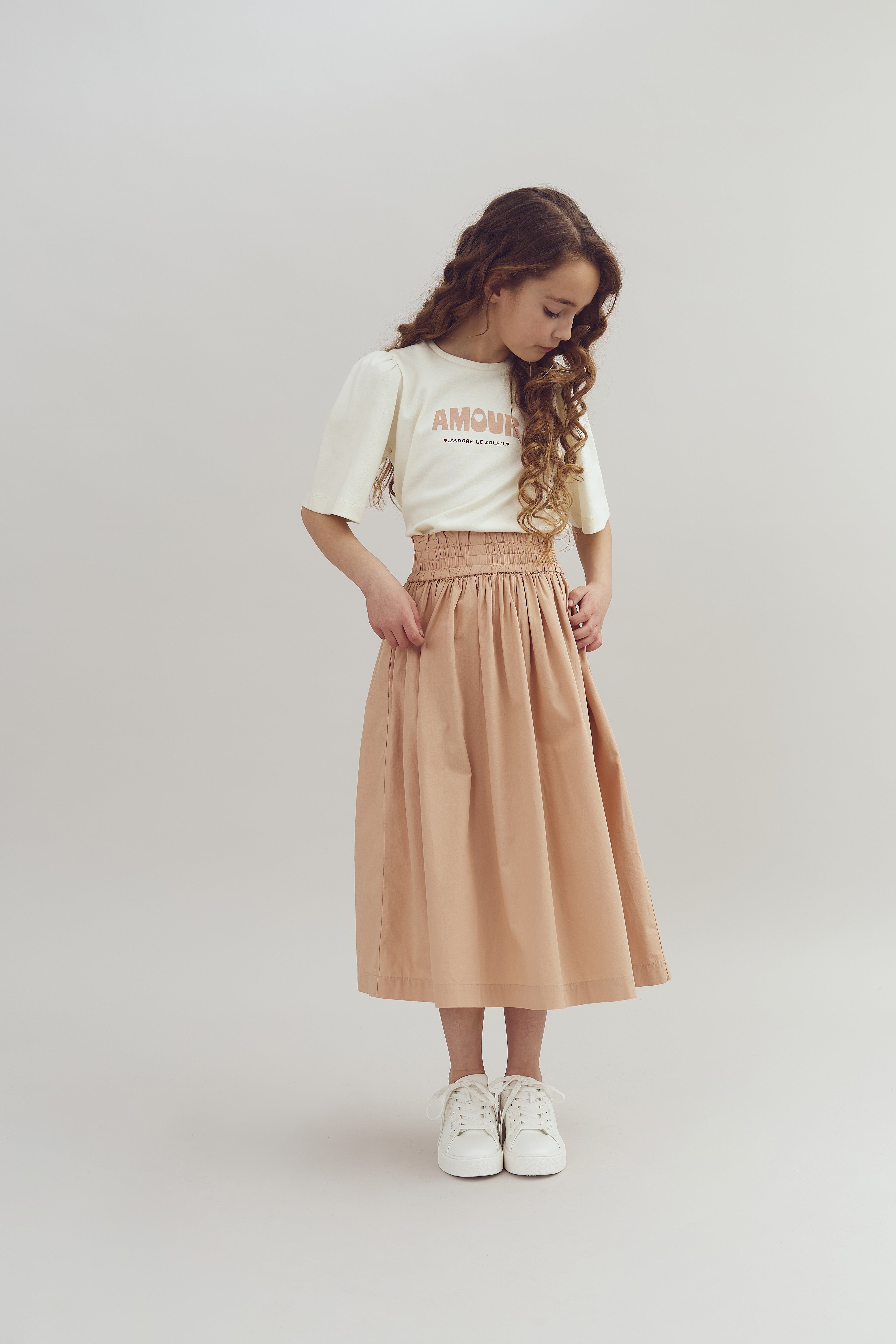 A young girl looks down at her beige skirt, standing against a plain background. She wears a white shirt with the text "AMOUR J'adore ce Soleil," paired with white sneakers.