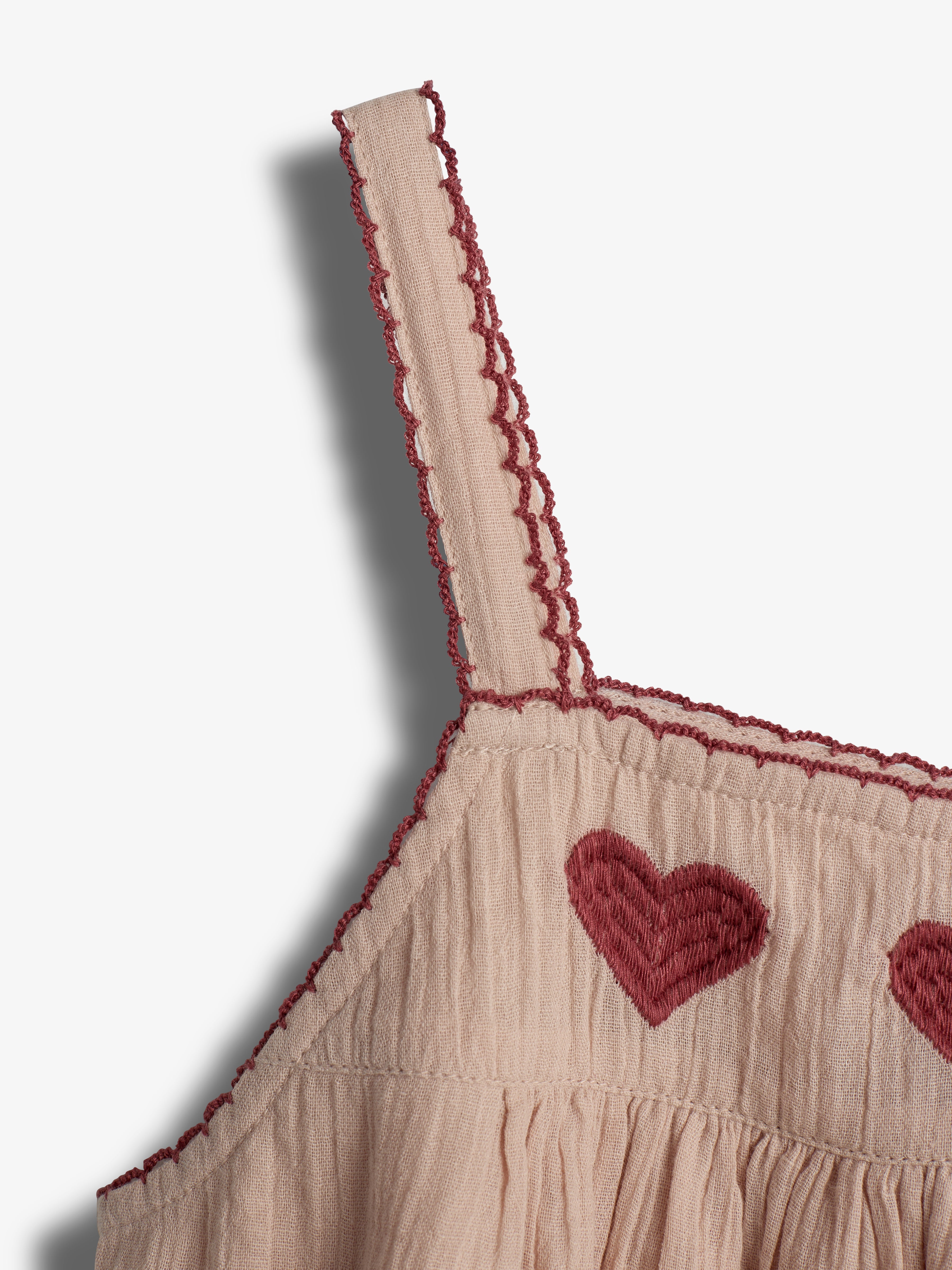 A beige dress strap adorned with red scalloped embroidery. Below, the dress features embroidered red hearts and a textured fabric. The background is plain white, creating a simple, elegant look.