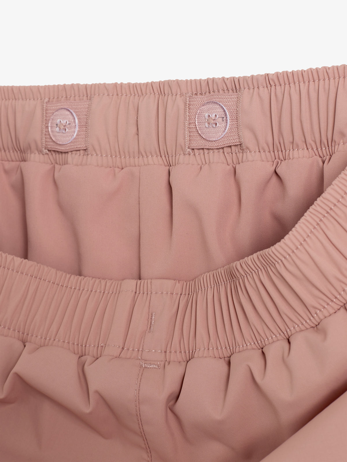 A pair of pink elastic waist shorts with adjustable buttons is displayed, highlighting the waistband design and stitching. The background is white, focusing attention on the shorts' details.