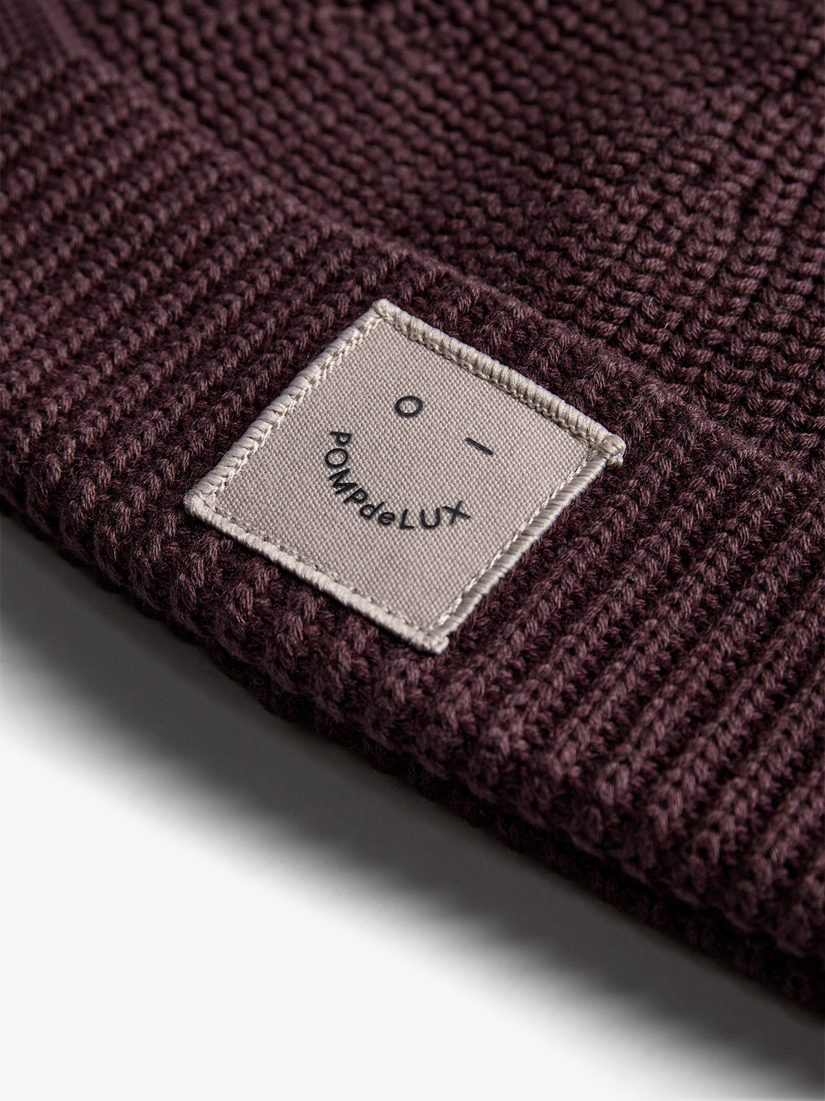 A close-up of a knitted burgundy fabric, featuring a sewn-on beige label with a simple face and text: "POMPdeLUX." The background is softly lit, emphasizing texture.