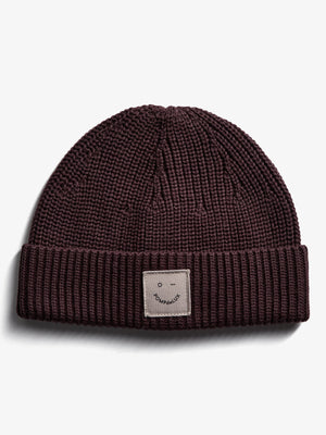 A knitted beanie resting flat on a white surface features a visible patch with a smiley face and "POMPdeLUX" text on the folded brim.