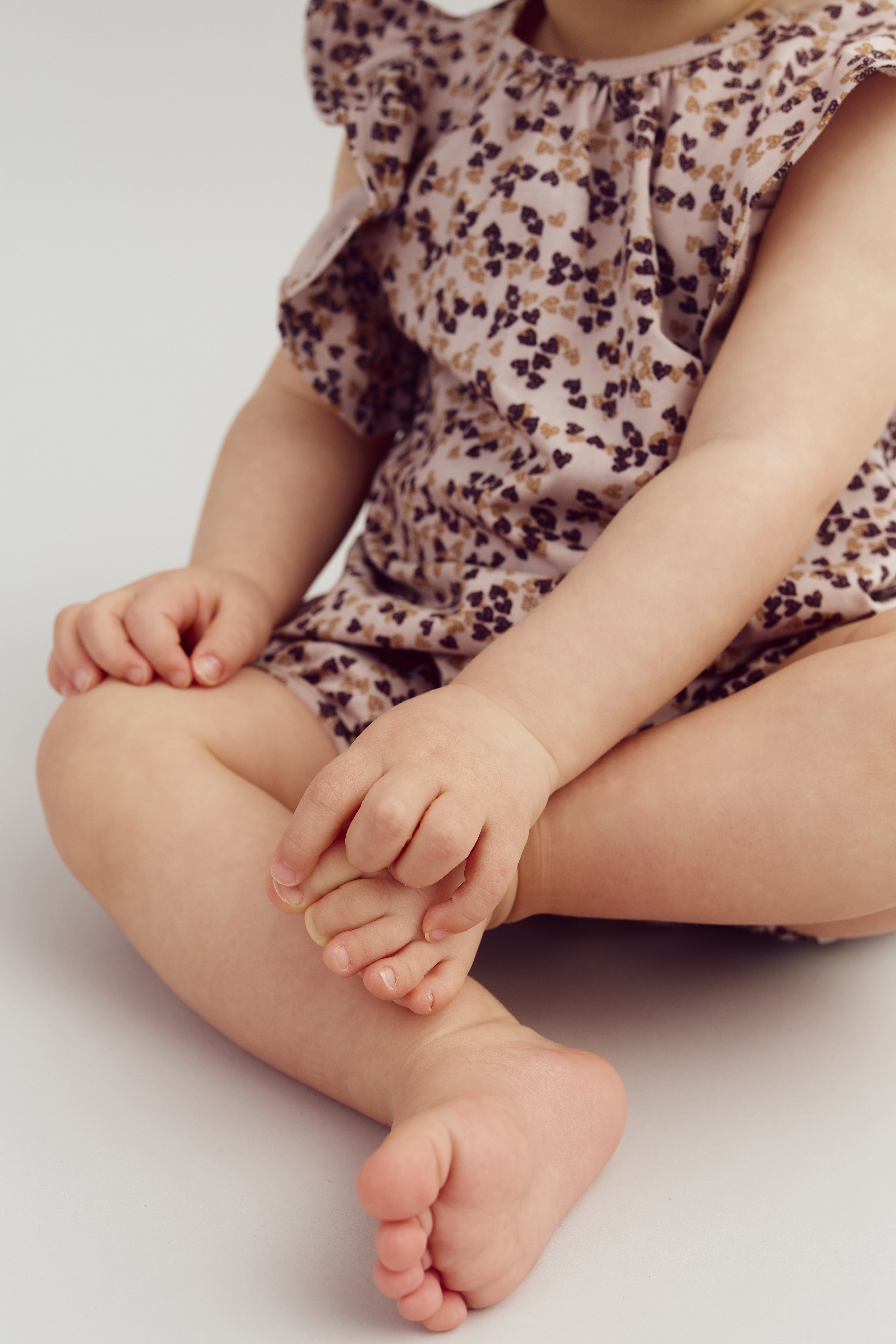 A baby is sitting cross-legged, wearing a floral romper, and holding its foot, set against a plain, light-colored background.