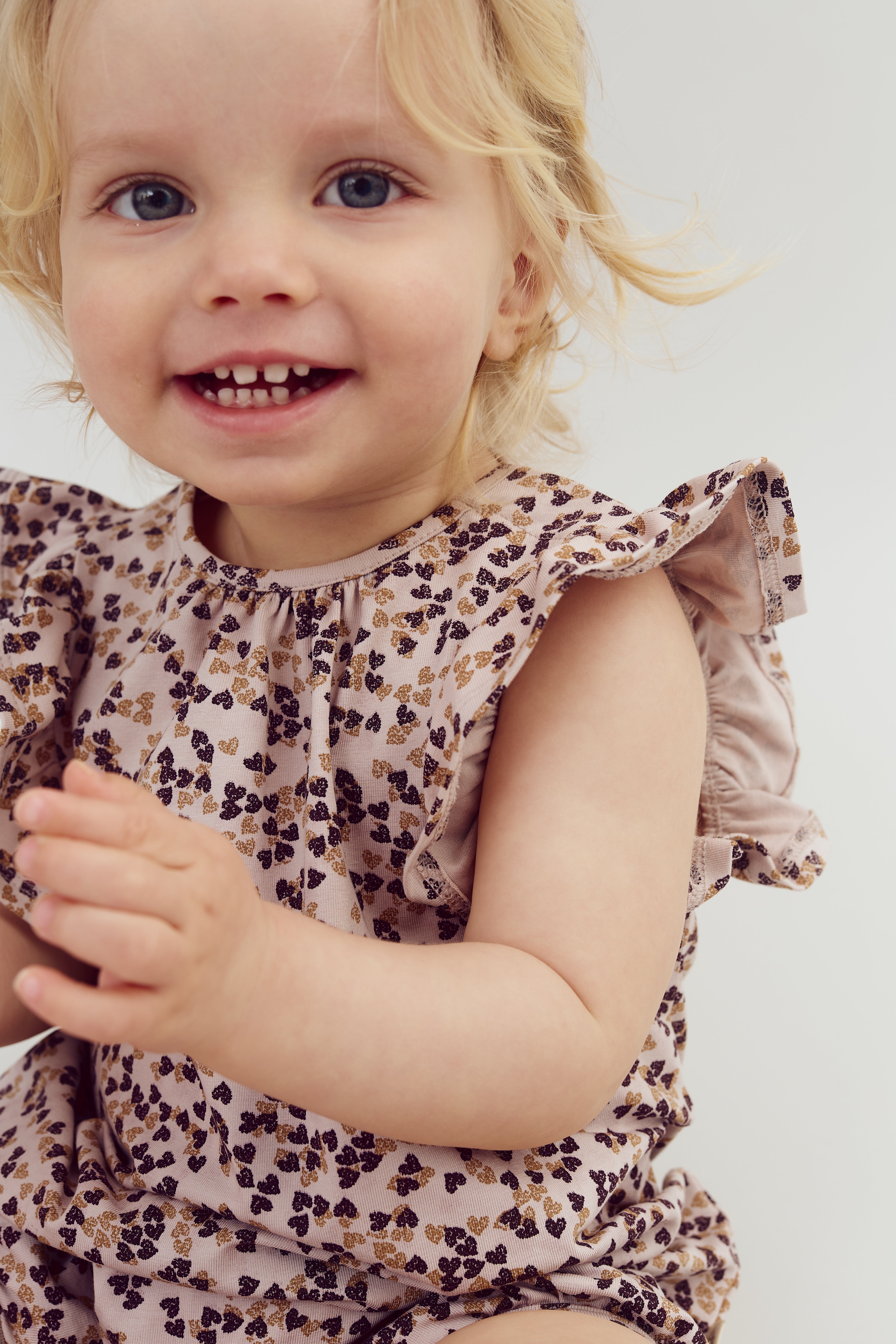 A young child with blonde hair smiles and claps, wearing a patterned ruffled outfit. The background is a plain light color, creating a joyful and simple atmosphere.