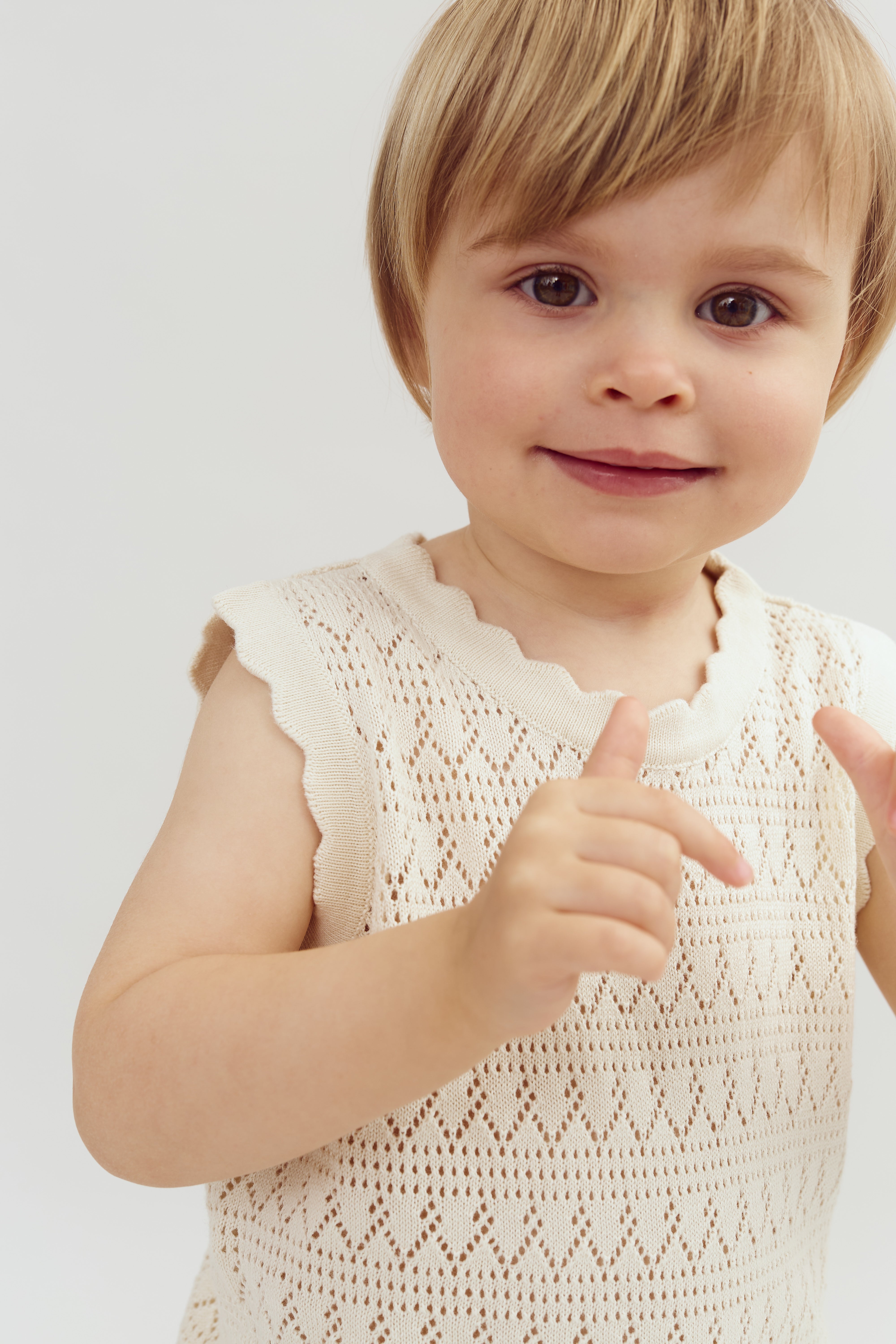 A young child with light hair is wearing a sleeveless, knitted cream top, smiling and gesturing playfully against a plain white background.