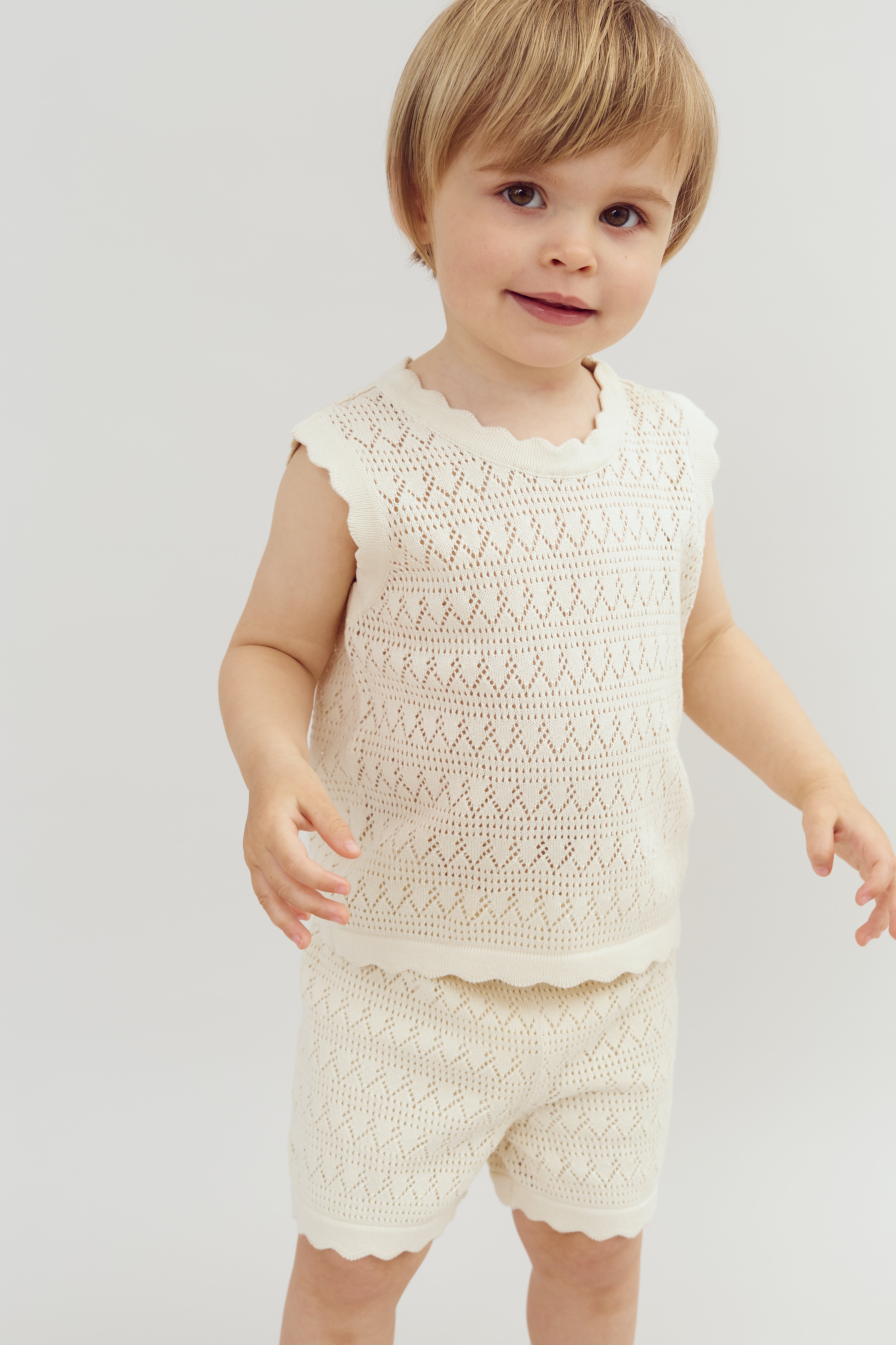 A young child, wearing a knitted cream top and shorts set, stands smiling with outstretched arms against a plain white background.