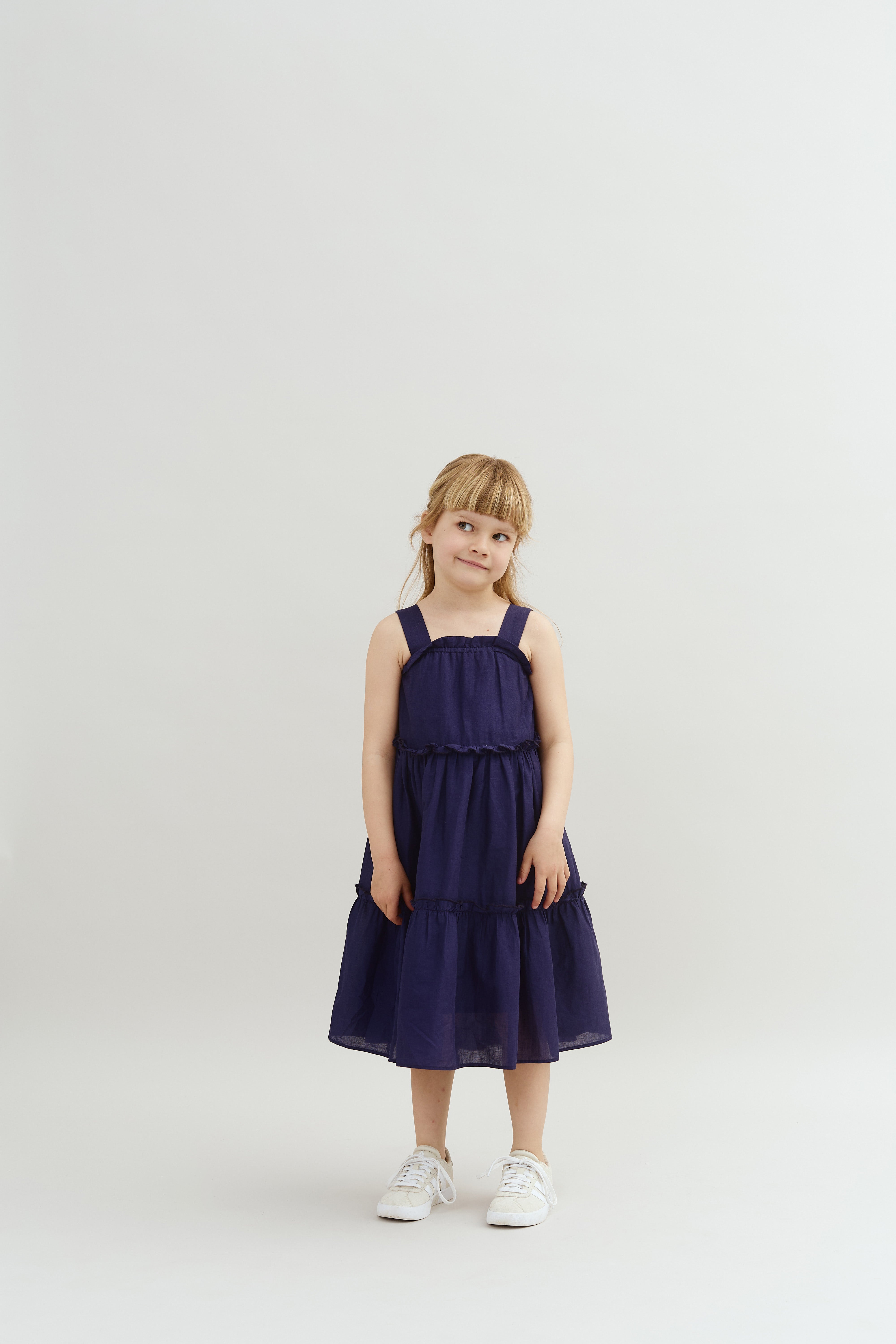 A young girl stands, wearing a navy dress and white sneakers, in a minimalist studio with a plain, light background.