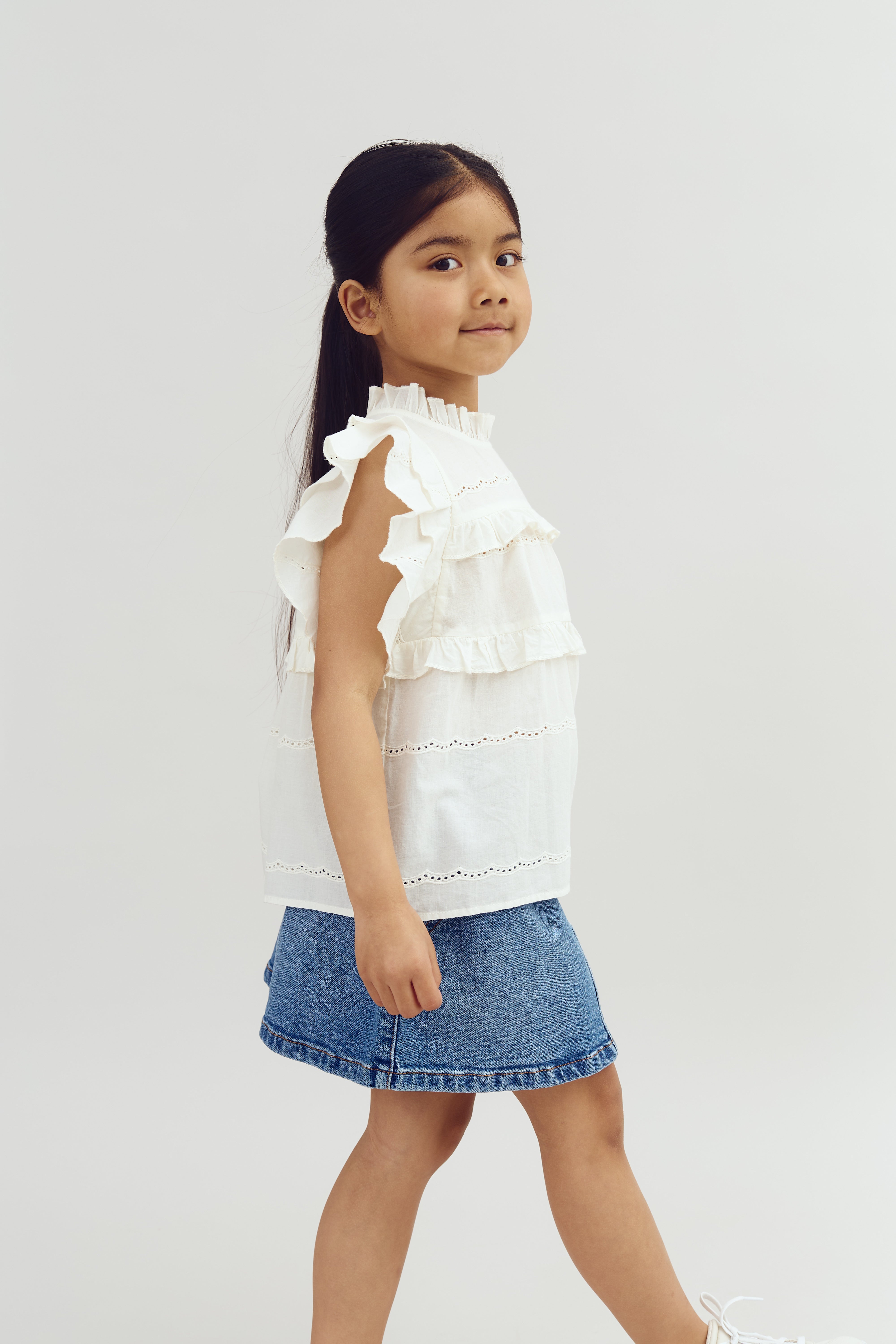 A young girl walks confidently, wearing a frilly white top and denim skirt, against a plain white background.