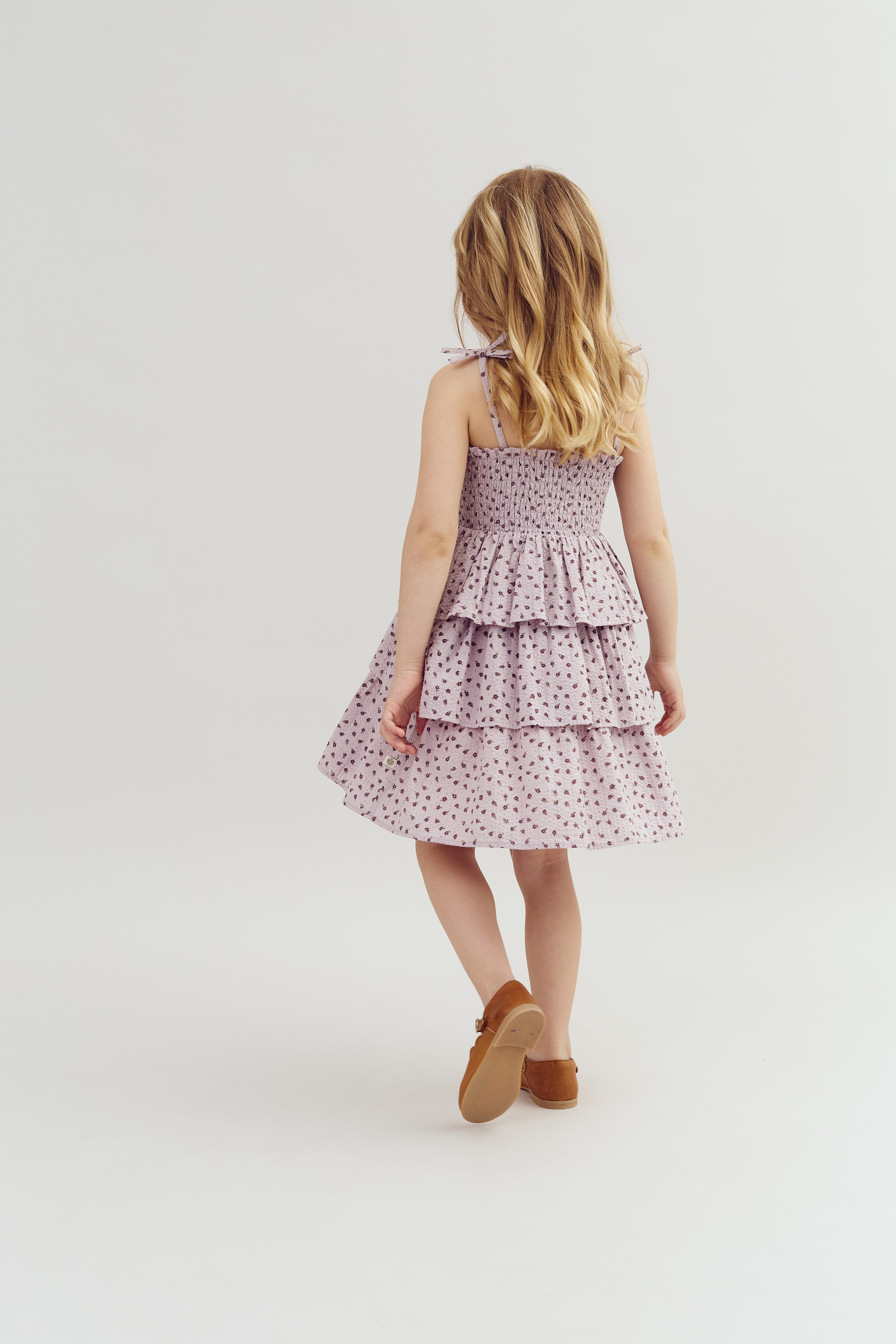 A young girl, with long blonde hair, walks away wearing a tiered, floral-patterned sundress and brown shoes against a plain, light background.