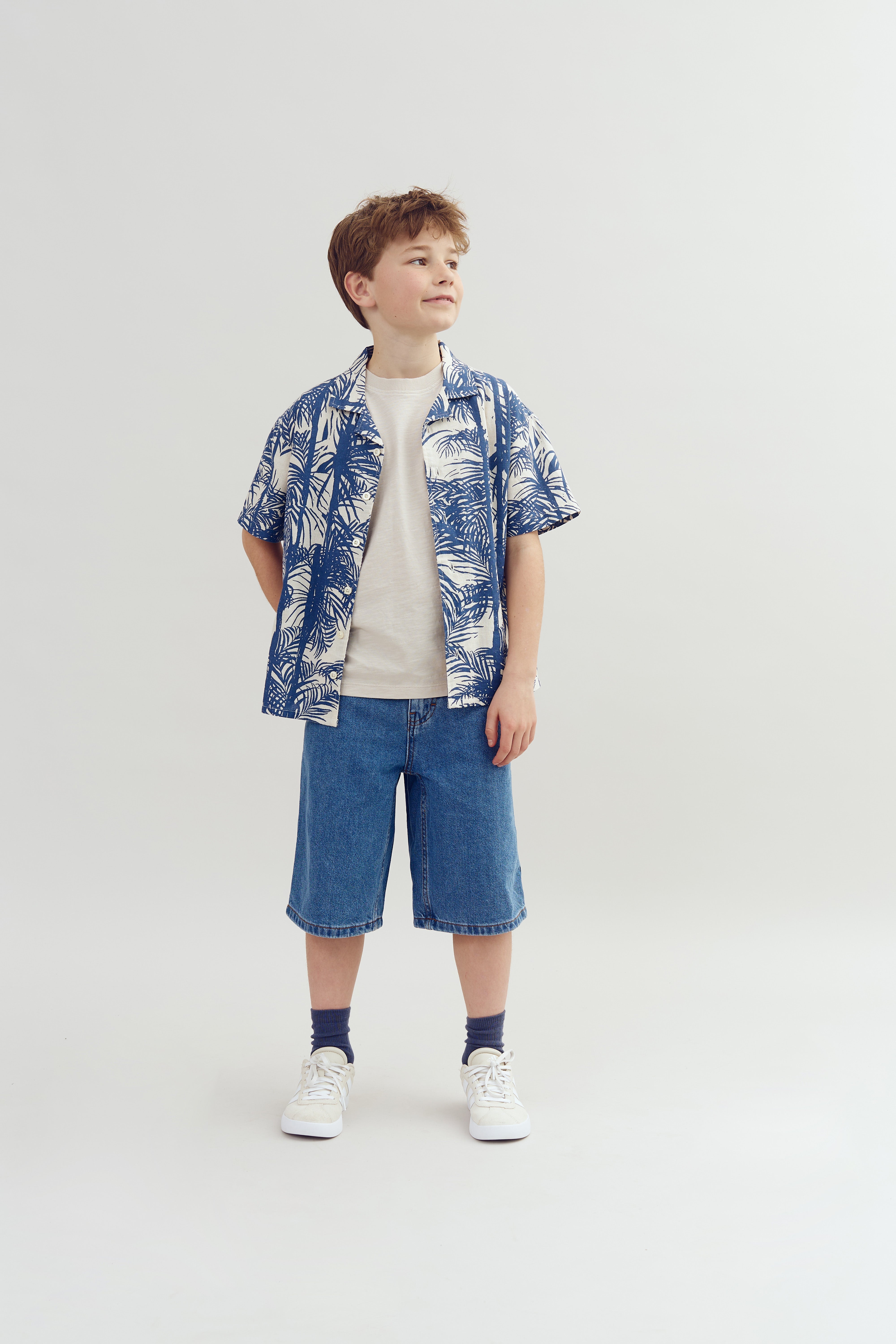 A young person stands confidently, wearing a blue patterned shirt, beige t-shirt, denim shorts, and white sneakers. The background is plain and neutral, enhancing focus on the outfit.