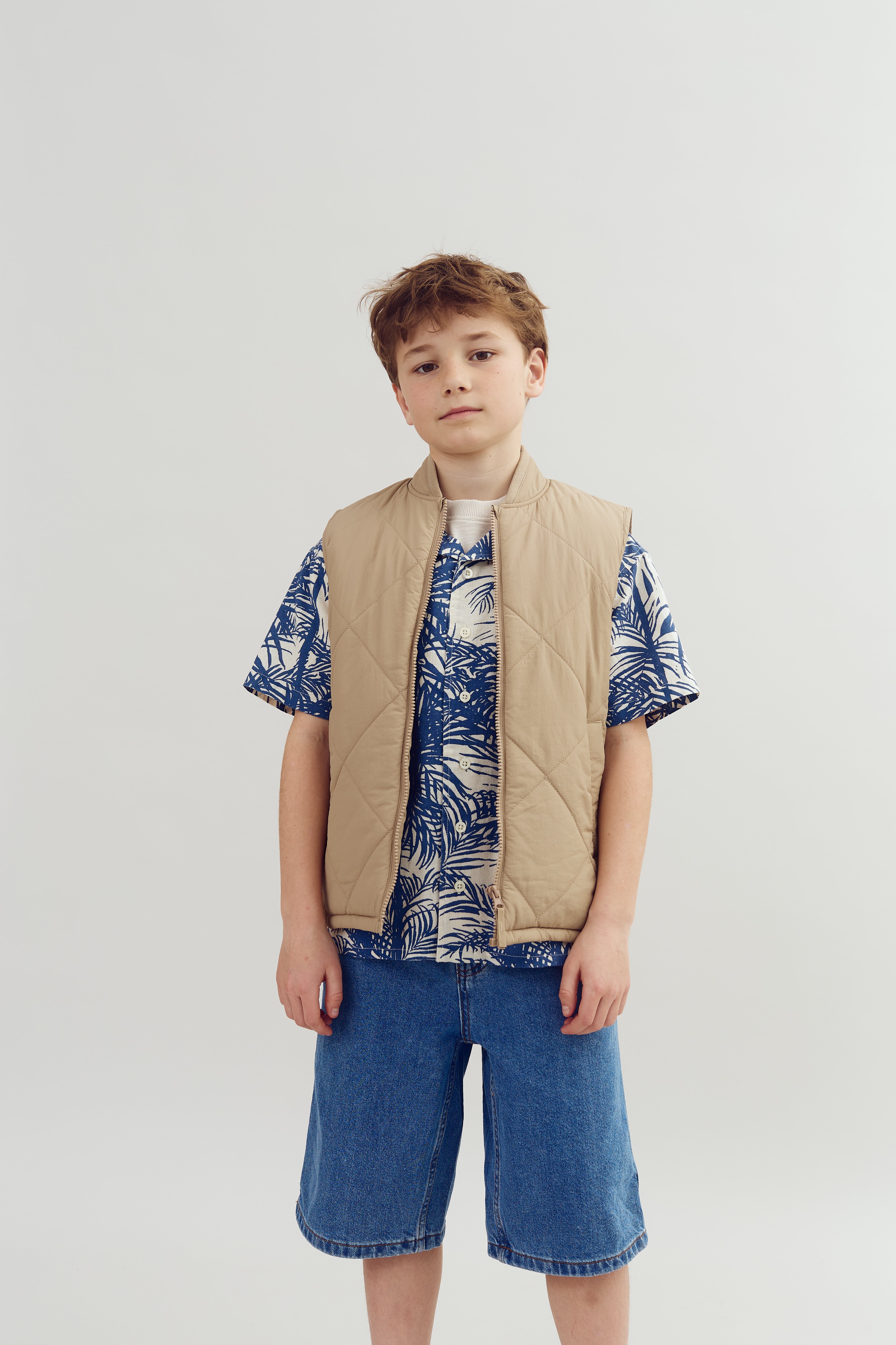 A young person stands, wearing a beige quilted vest over a blue leaf-patterned shirt and denim shorts, set against a plain white background.