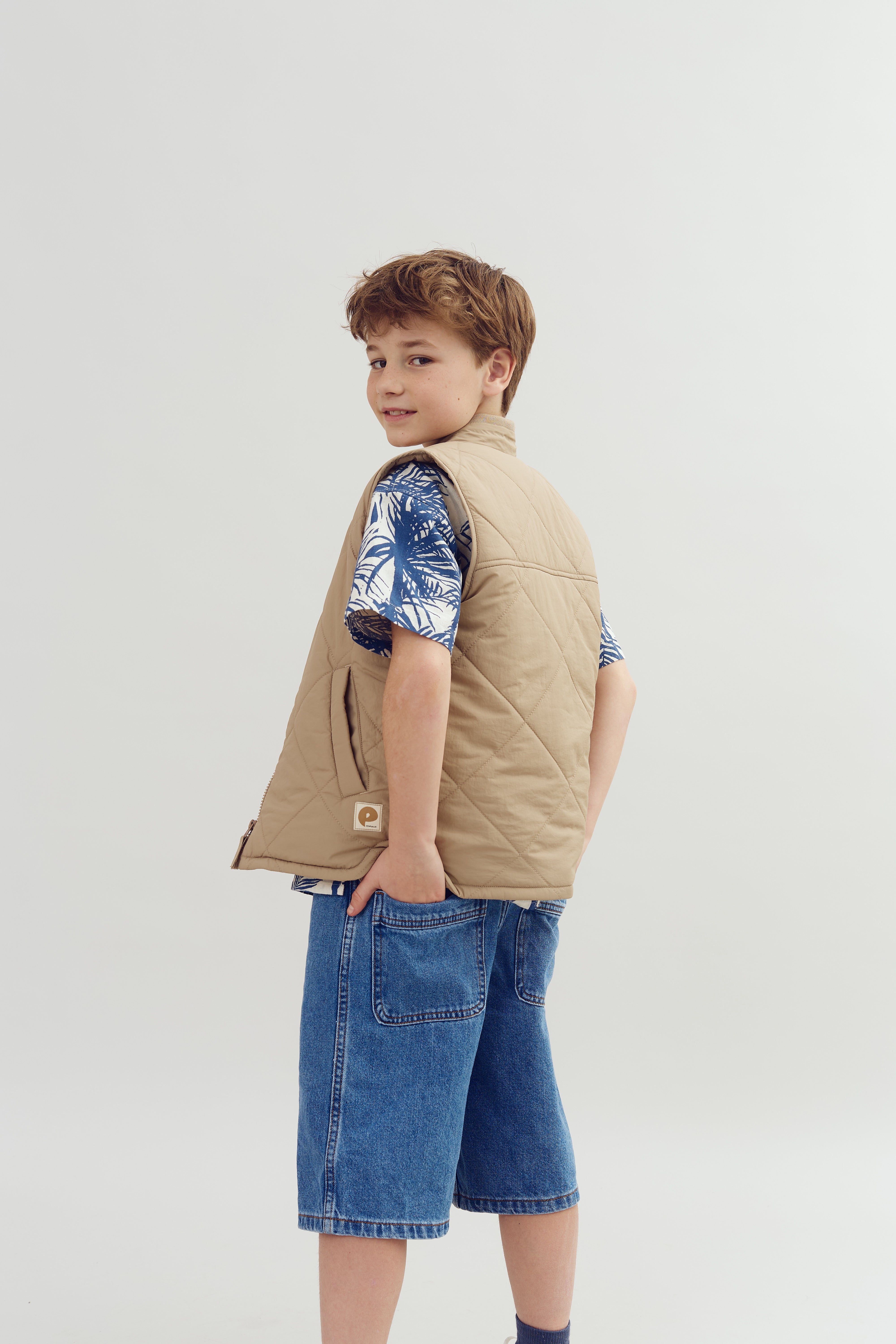 A child wearing a tan quilted vest and blue tropical shirt stands with hands in pocket, looking back. They are in blue jean shorts against a plain, light background.